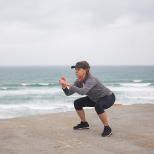 Mature woman in chair yoga pose on a concrete pier by the ocean wearing Herron Apparel leggings and eco-friendly jacket.